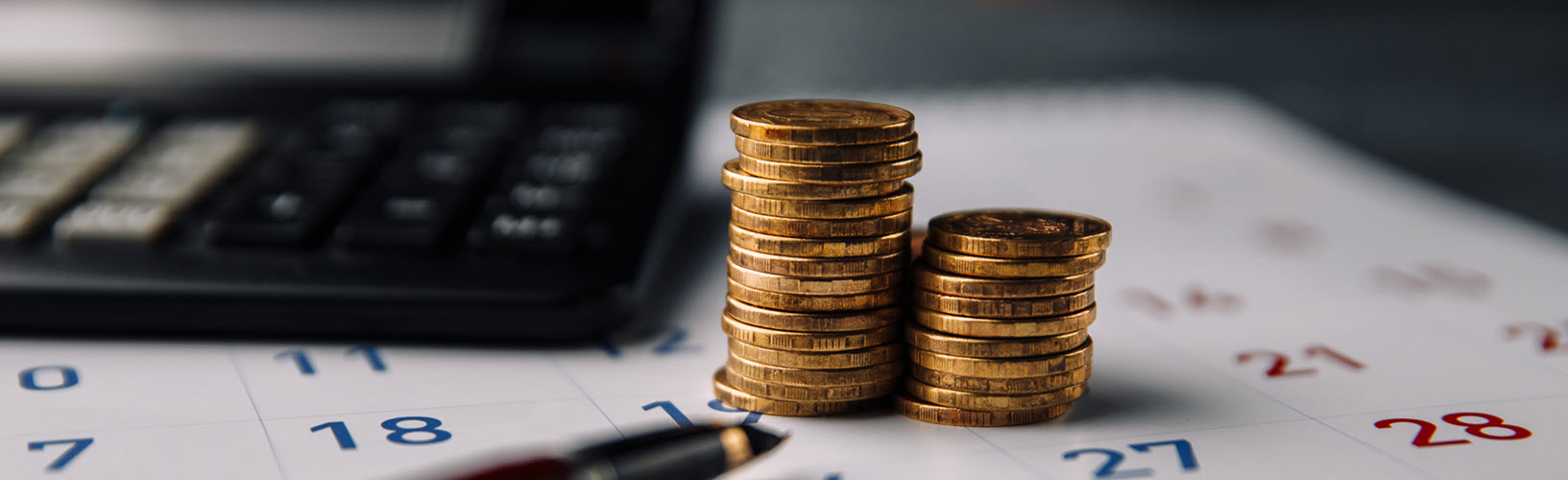 Two stacks of gold coins on top of a desk calendar next to a calculator and with a pen in front.