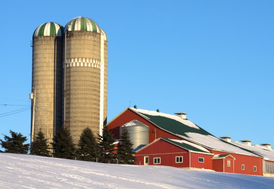 a snow covered farm with a red barn and hay bales