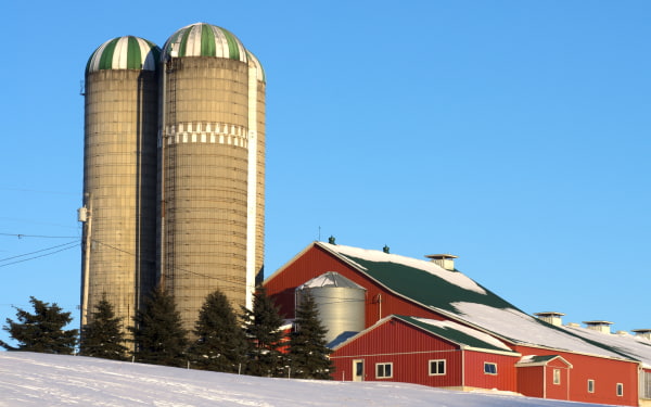 a snow covered farm with a red barn and hay bales
