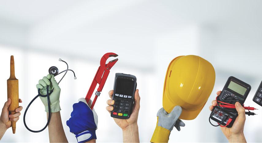 Hands holding up tools of multiple professions against a white background: rolling pin, stethoscope, wrench, card reader, hardhat, and a multimeter.