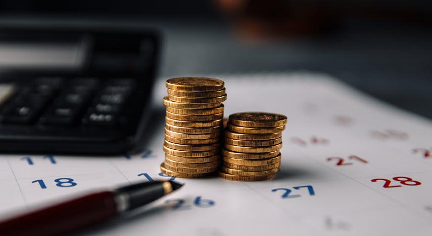 Two stacks of gold coins on top of a desk calendar next to a calculator and with a pen in front.