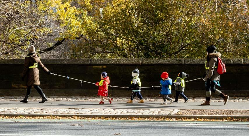 children walking in a line, holding a rope, between adults on each end of the rope